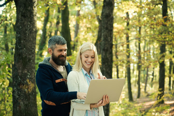 Autumn couple using notebook. Autumn outdoor portrait of beautiful happy girl and bearded man walking in park or forest. Fashion autumn portrait woman and man with laptop on nature background.