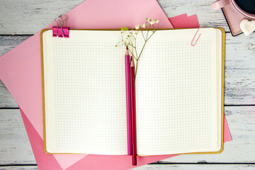 An open notebook in a cage on pink album sheets, next to two red pencils, a pink coffee chicken, an old pocket watch on of the wooden table. A flat lay style composition, a top view 