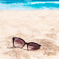Fototapeta na wymiar Old lost abandoned sunglasses on beach sand with turquoise Caribbean ocean in background in bright summer day. Tropical summer vacation concept 