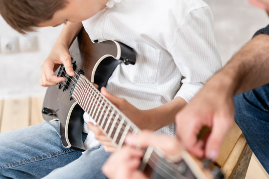 Young boy moves his hand along guitar strings while his father helps boy to take chords