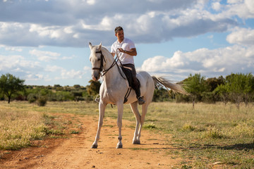 Young guy in casual outfit riding white horse on meadow a sunny day 