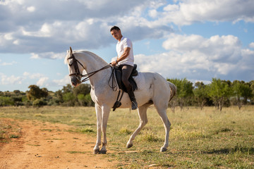 Young guy in casual outfit riding white horse on meadow a sunny day 