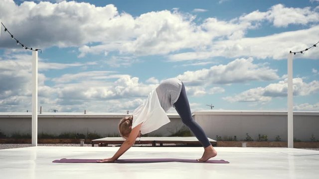 Flexible woman does yoga in slow motion in background of white clouds and bright blue sky. Girl changes yoga pose. Downward facing dog pose - Adho Mukha Svanasana. One Legged Down Dog pose