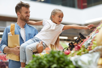 Portrait of cute little girl reaching for vegetables while enjoying grocery shopping with dad at...