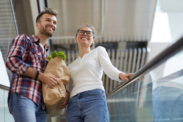 Low angle portrait of contemporary young couple riding escalator in shopping mall and carrying...
