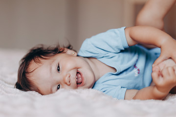 cute laughing one year old girl lying on bed and looking at camera touch her feet