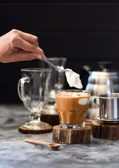 Woman hand pouring whipped coconut cream into blended Vietnamese drip coffee in glass on dark background copy space