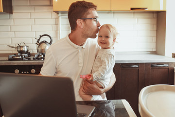 Happy young father working on laptop at home with little baby daughter. Multitasking.