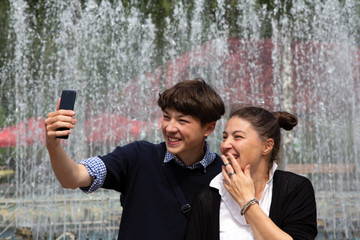 Middle-aged woman, mother, and teenager, son take a selfie at the fountain in the city park. Family resting together on the weekend.