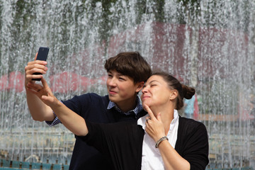 Middle-aged woman, mother, and teenager, son take a selfie at the fountain in the city park. Family resting together on the weekend.