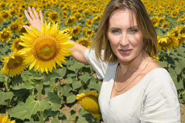 Close up portrait of a pretty woman looks at the camera as she shows a field of sunflowers behind her. She has a large sunflower by her side