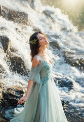 Fototapeta na wymiar charming girl with light hair in long blue turquoise delicate dress with deep neckline and open shoulders near fast flow of water, birth of mermaid in waterfall in bright light of warm yellow sun
