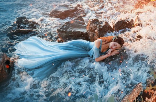 charming sea princess resting on wet stones, girl in blue long tender dress sweetly sleeps in cold water of waterfall with hands under head, sweet red mermaid with kind face in rays of warm sun