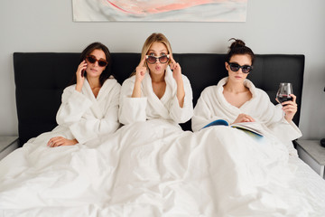 Three young beautifu women in white bathrobes and sunglasses lying in bed in modern cozy hotel. Pretty girl amazedly looking in camera while girls near talking on cellphone and reading book