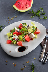 Summer salad with watermelon and feta cheese salad in a white plate. Brown tablecloth, spoon, fork on the grey background. Top view