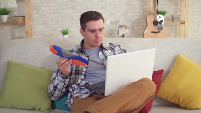 man sitting at a table uses a laptop and holds orthopedic insoles in hand