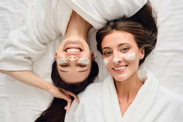 Two young gorgeous smiling women with dark hair in white bathrobes lying in bed with cosmetic mask...