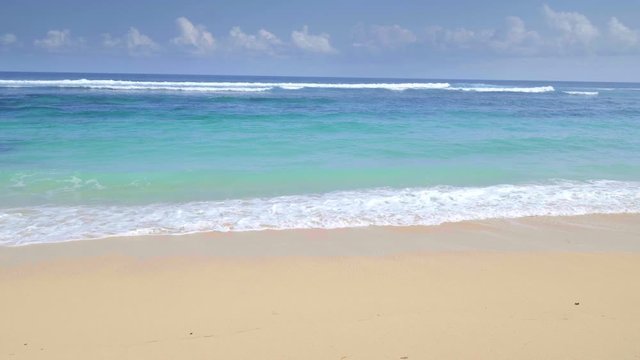 A panoramic shot of tropical sandy beach and rocks with colorful blue water and waves filmed in slow motion