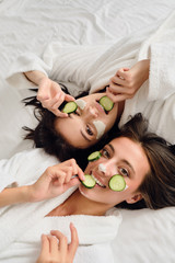 Two young attractive women with dark hair in white bathrobes with cosmetic mask and slices of cucumber on faces lying in bed dreamily looking in camera together in modern cozy hotel