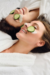 Two young attractive smiling women with dark hair in white bathrobes with cosmetic mask and slices...