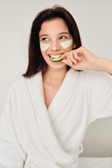 Young beautiful smiling woman in white bathrobe with cosmetic mask on face sitting on bed eating slice of cucumber happily looking aside in modern cozy hotel