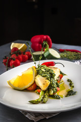 Baked Halibut fish steak with lemon, carrot and sauce salsa verde. White plate, thyme, spoon, fork on the grey background