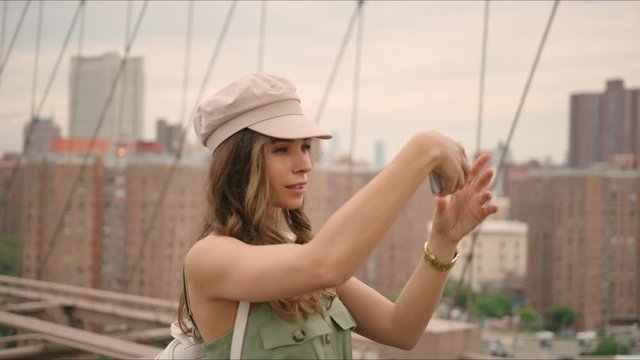 French tourist walks by Brooklyn Bridge and takes photos on a phone, first time in New York, visiting famous destinations, moment from life, making content. 