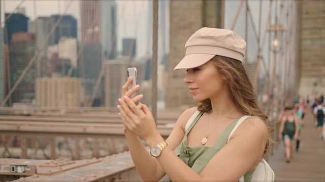 French tourist walks by Brooklyn Bridge and takes photos on a phone, first time in New York, visiting famous destinations, moment from life, making content. 