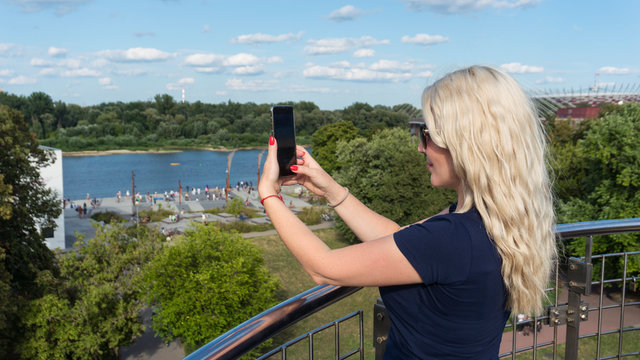 Back view of a woman taking photograph with a smart phone camera. Pretty female tourist is taking pictures of the River.