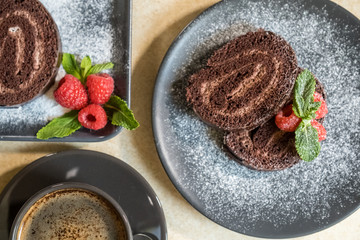 Chocolate swiss roll with jam decorated with raspberries on a plate and black coffee on the table. Selective focus.