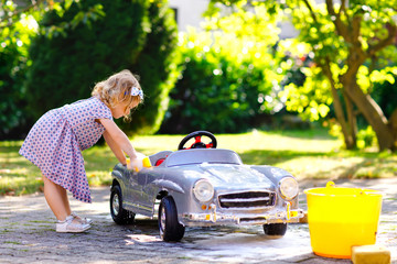 Cute gorgeous toddler girl washing big old toy car in summer garden, outdoors. Happy healthy little child cleaning car with soap and water, having fun with splashing and playing with sponge.