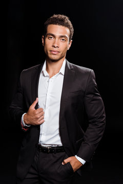 handsome mixed race man touching suit while standing with hand in pocket isolated on black
