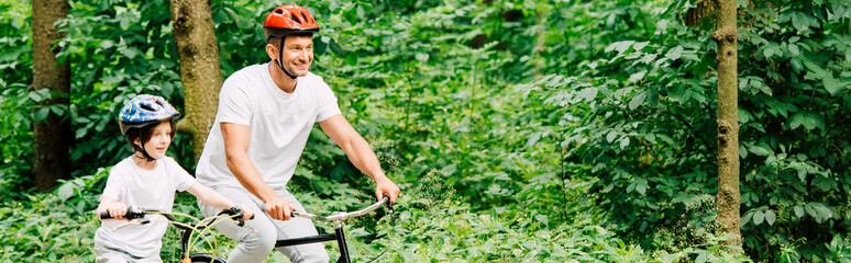 panoramic shot of father and son smiling while riding bicycles in forest