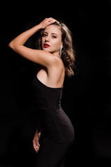 elegant young woman touching hair while standing in dress isolated on black