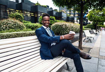Obraz premium African American businessman wearing blue suit sits on the bench and drinks coffee near office. Business concept
