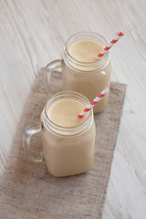 Glass jar mugs filled with peanut butter banana smoothie on a white wooden background, low angle view. Close-up.