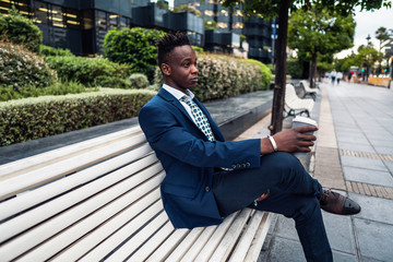 Obraz premium African American businessman wearing blue suit sits on the bench and drinks coffee near office. Business concept