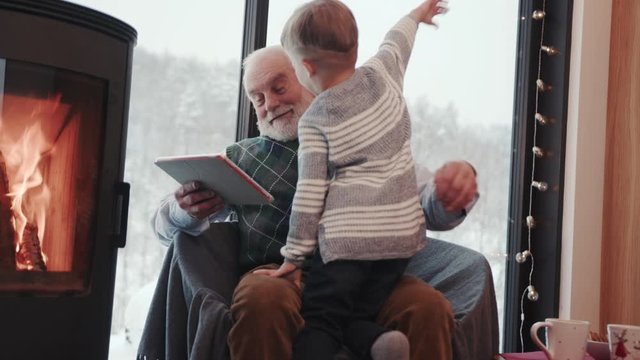 Slow motion grandfather and grandson using digital tablet for surfing internet and playing game near the fireplace at home grandpa adult grandchild child childhood communication computer