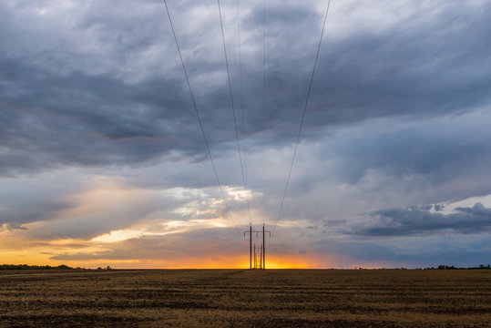 A row of high voltage electrical power lines cling to the horizon in an otherwise vast, wide open rural landscape on sunset with clouds