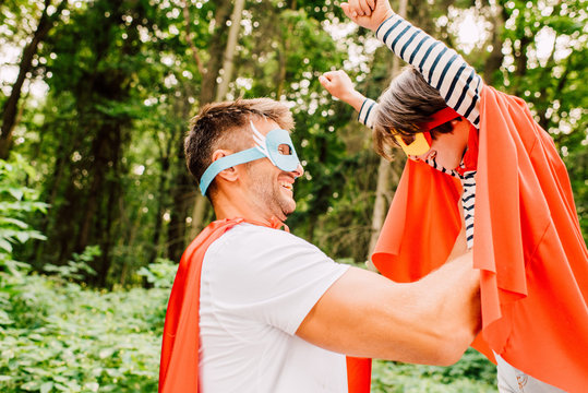 side view of father in superhero costume holding son in hands while kid laughing and looking at dad