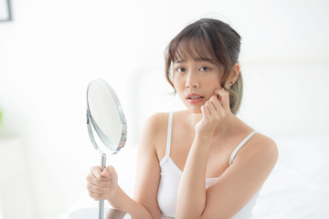 Beautiful young asian woman looking the mirror with acne problem at bedroom, trouble of beauty on face, zit treatment, asia girl is pimple having worry and displeased, skincare and healthy concept.