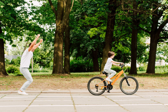 Side View Of Happy Father Cheering Son While Kid Riding Bicycle