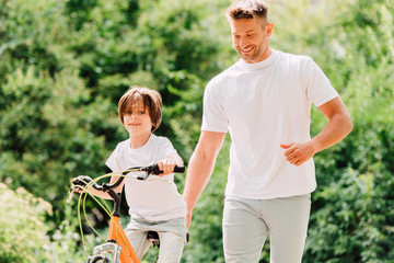 son riding bicycle and father walking next to kid and holding sit of bike