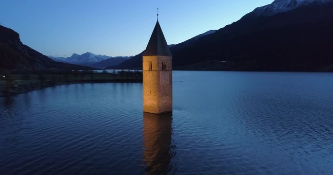 Aerial: Slow Pan of the Lake Reschen Bell Tower at Night in front of Snow Capped Mountains - South Tyrol, Italy