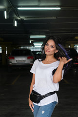 Fototapeta na wymiar Cool carefree brunette with colored strands of hair posing in underground parking. On girl modern summer outfit in style of casual. White T-shirt, jeans and waist bag. Freedom, independence, lifesty
