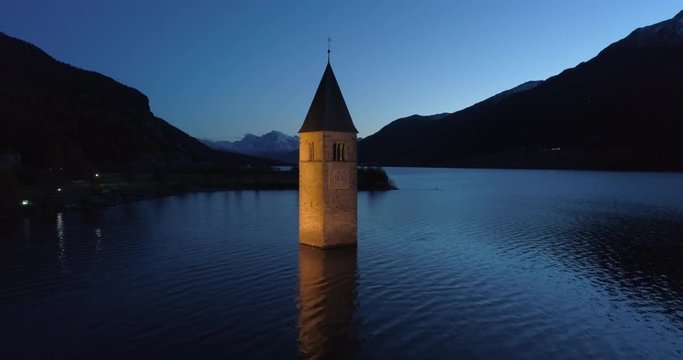 Rising Aerial: Illuminated Bell Tower of Lake Reschen and Distant Hills and Mountains at night - South Tyrol, Italy