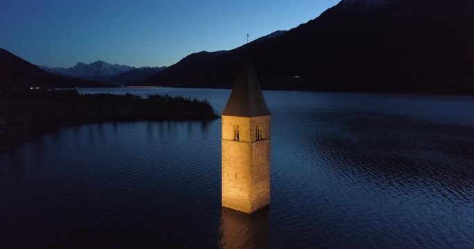 High Elevation Aerial: The Bell Tower of Lake Reschen at Night with Distant Lights, Hills, and Mountains - South Tyrol, Italy
