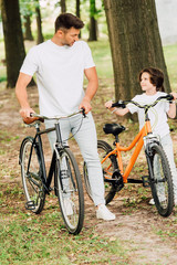 Fototapeta premium full length view of father and son walking in park with bicycles and looking at each other