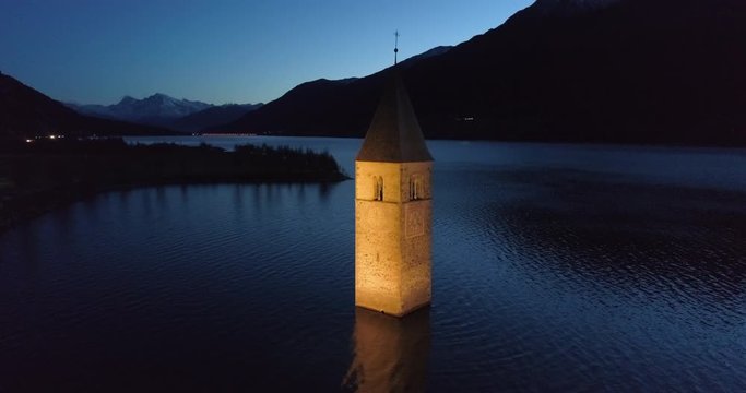 Aerial: Bell Tower of Lake Reschen Reflected in the Water at Night Before Hills and Mountains - South Tyrol, Italy