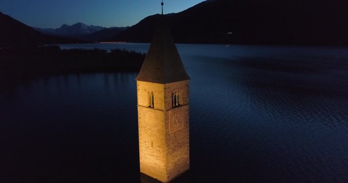 Aerial Pan: The Illuminated Bell Tower of Lake Reschen at Night With Dark Hills and Mountains - South Tyrol, Italy